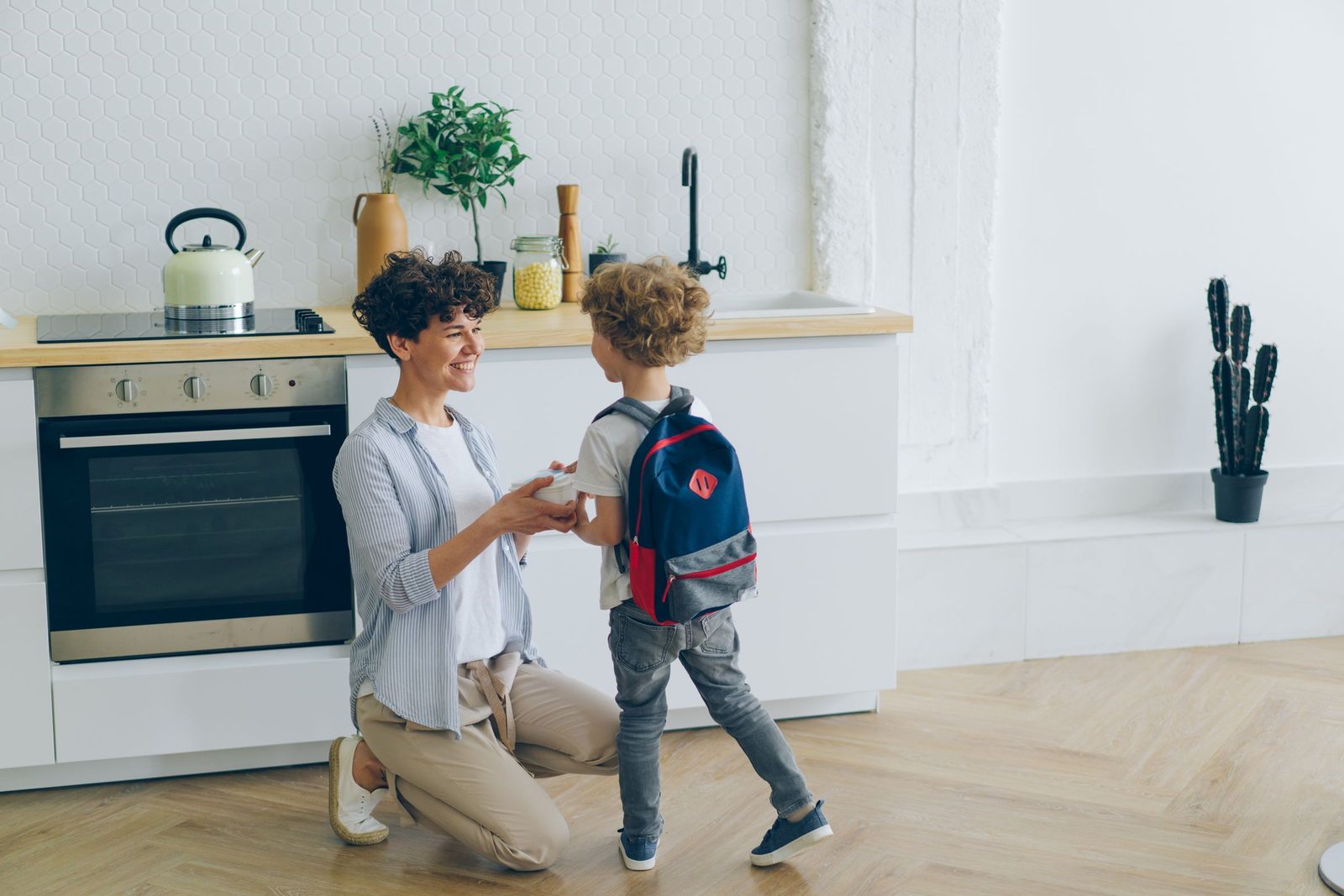 A smiling mother kneeling in a bright kitchen as her young son, wearing a backpack, stands beside her